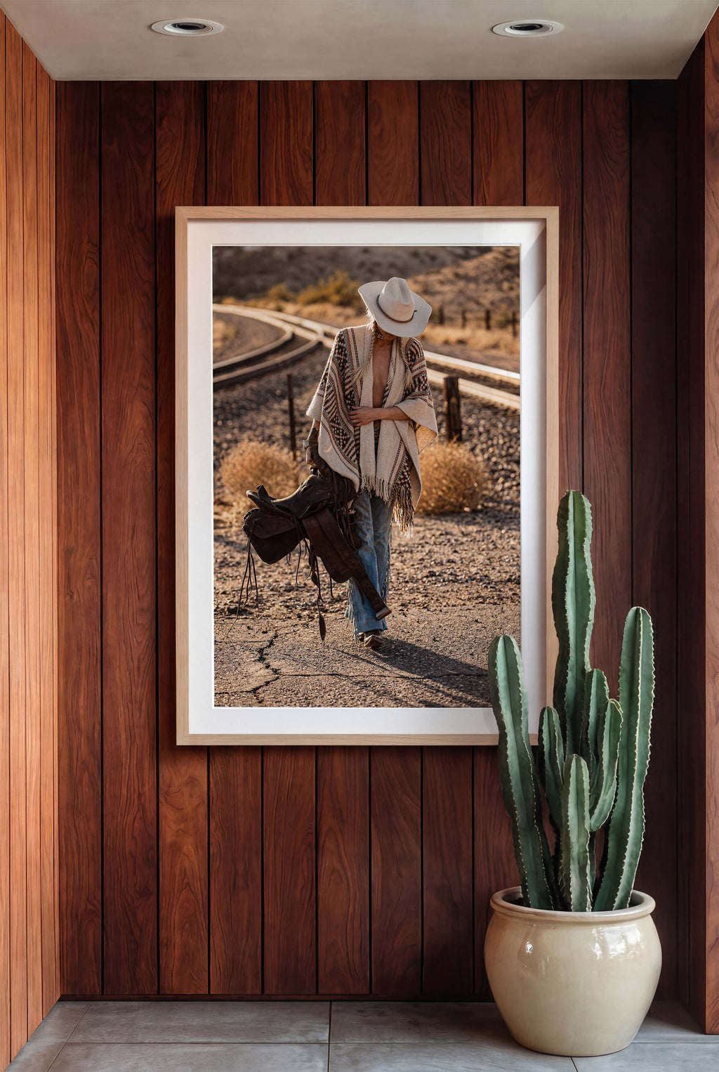 Framed photograph of a cowgirl in a desert landscape on a wooden wall with a cactus plant below.