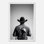 Black and white photograph of a rodeo cowboy viewed from behind wearing a western hat with a numbered rodeo tag pinned to his shirt after competition.