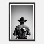Black and white photograph of a rodeo cowboy viewed from behind wearing a western hat with a numbered rodeo tag pinned to his shirt after competition.