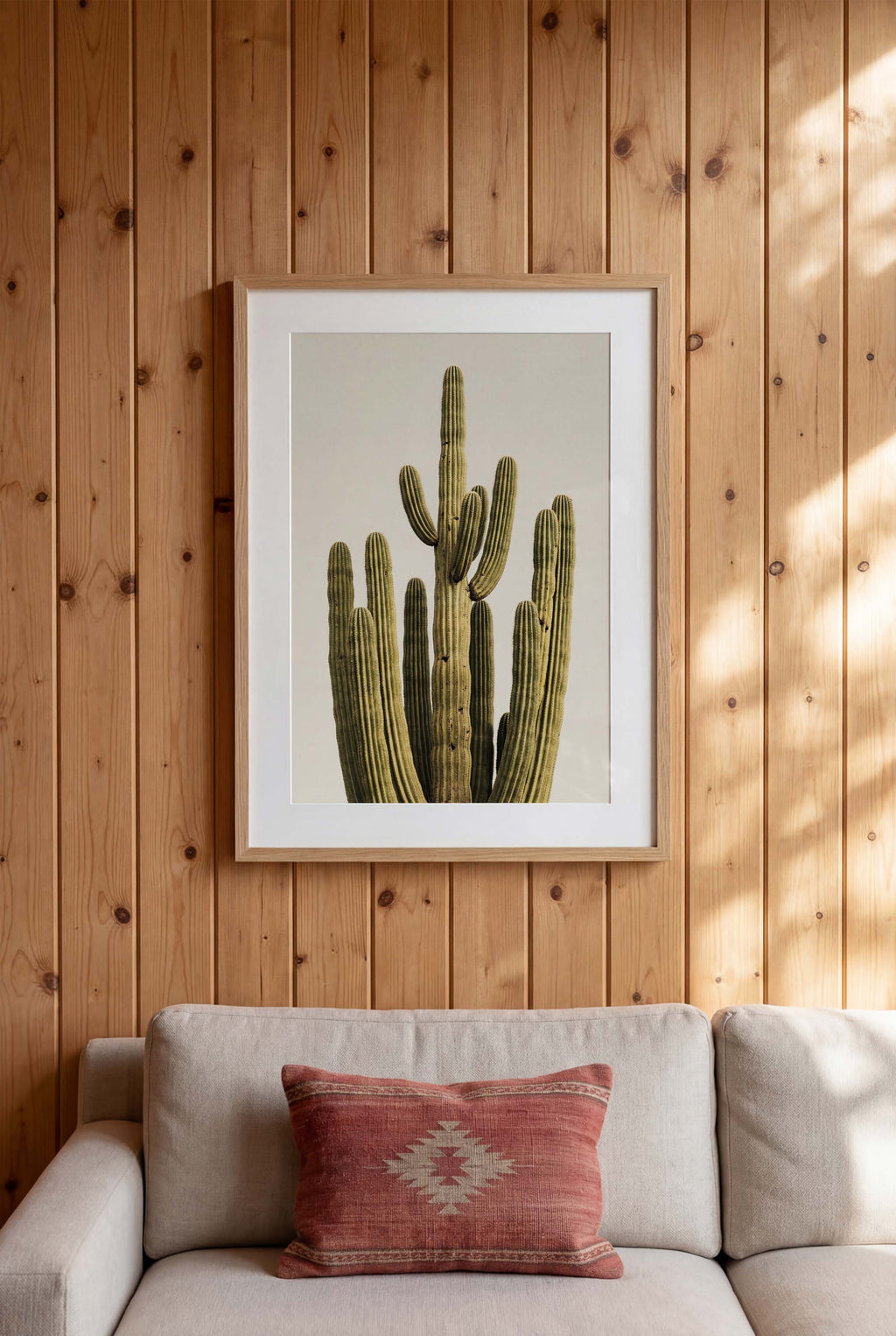 Framed picture of a cactus on a wooden wall above a beige sofa with a red pillow.