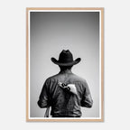 Black and white photograph of a rodeo cowboy viewed from behind wearing a western hat with a numbered rodeo tag pinned to his shirt after competition.