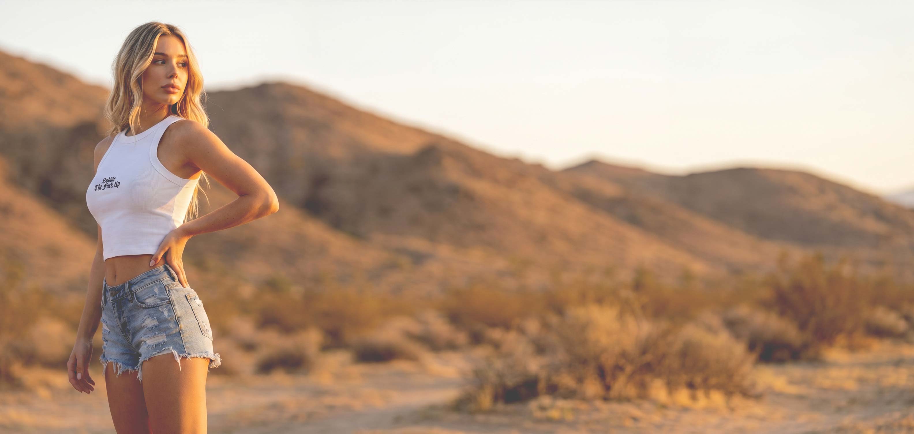Woman in a white tank top and denim shorts standing in a desert landscape with mountains in the background.