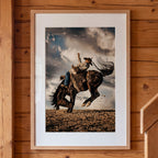 Framed artwork of a cowboy riding a bucking bronco against a cloudy sky, mounted on a wooden wall.
