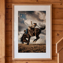 Framed artwork of a cowboy riding a bucking bronco against a cloudy sky, mounted on a wooden wall.