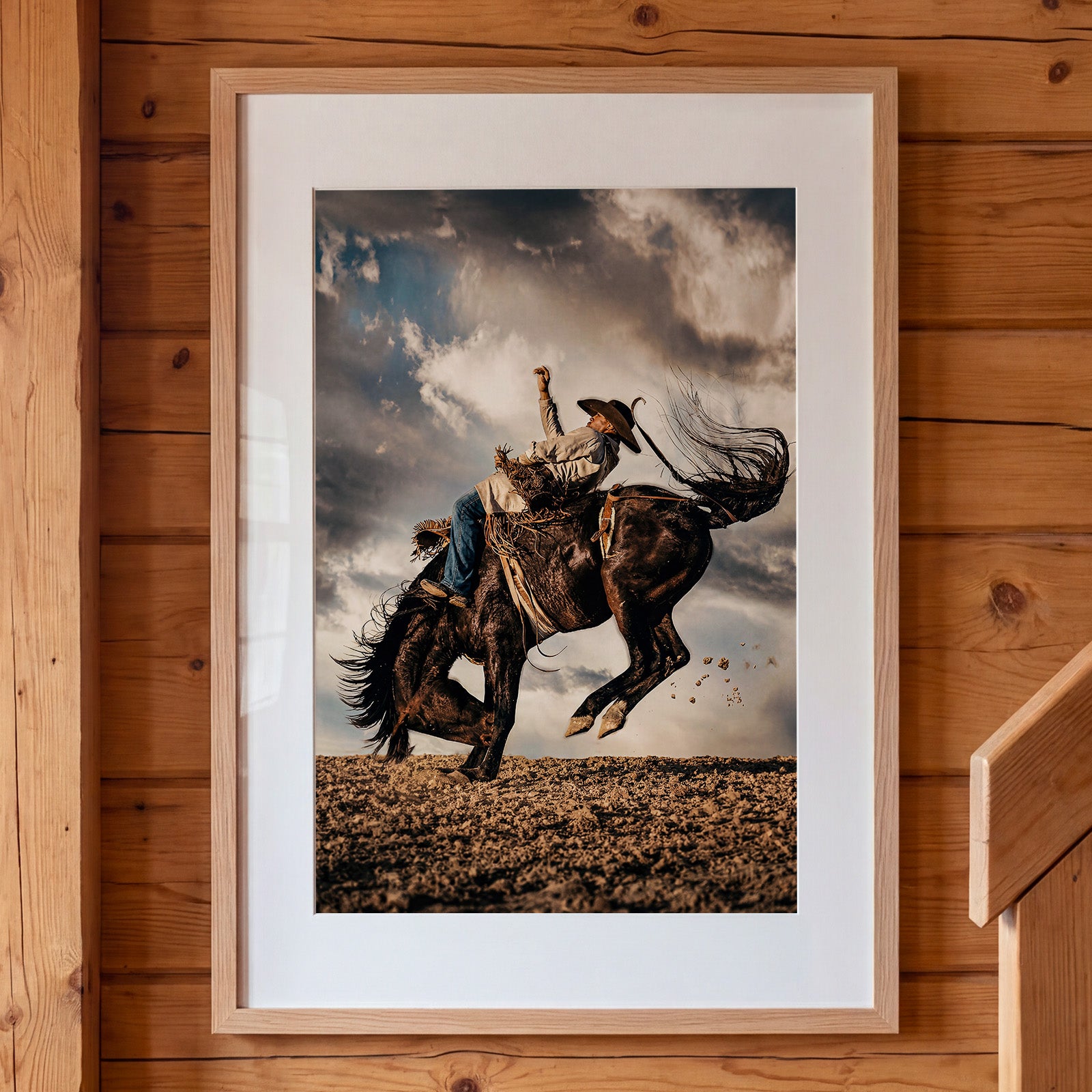Framed artwork of a cowboy riding a bucking bronco against a cloudy sky, mounted on a wooden wall.