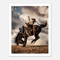 Cowboy riding a bucking horse at sunset with dust and dramatic sky, western rodeo wall art print